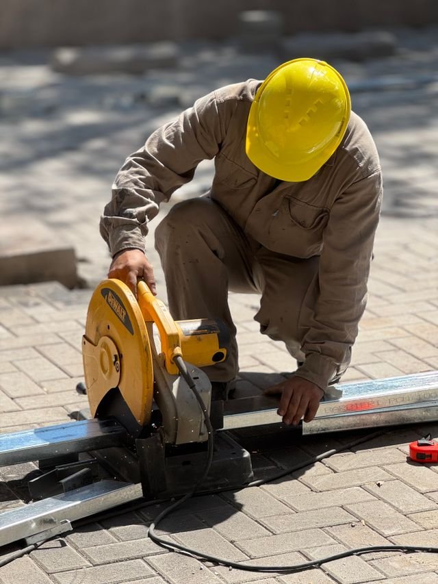 A construction worker wearing a yellow helmet cuts metal using a circular saw outdoors.